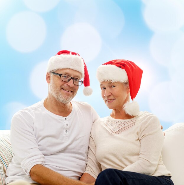 family, holidays, christmas, age and people concept - happy senior couple in santa helper hats sitting on sofa over blue lights background