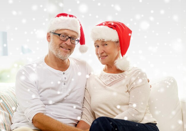 family, holidays, christmas, age and people concept - happy senior couple in santa helper hats sitting on sofa at home