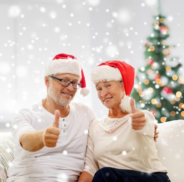 family, holidays, christmas, age and people concept - happy senior couple in santa helper hats sitting on sofa over living room and christmas tree background