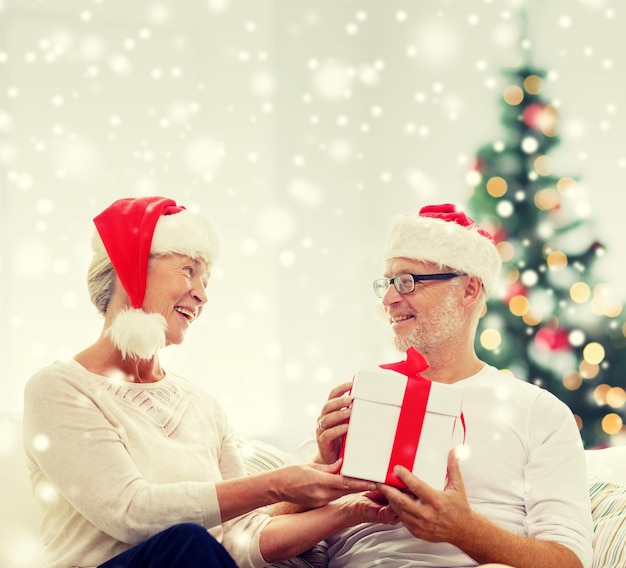 family, holidays, christmas, age and people concept - happy senior couple in santa helper hats with gift box at home