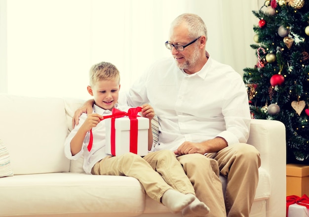 family, holidays, generation, christmas and people concept - smiling grandfather and grandson with gift box sitting on couch at home