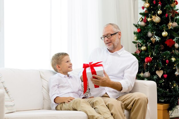 family, holidays, generation, christmas and people concept - smiling grandfather and grandson with gift box sitting on couch at home