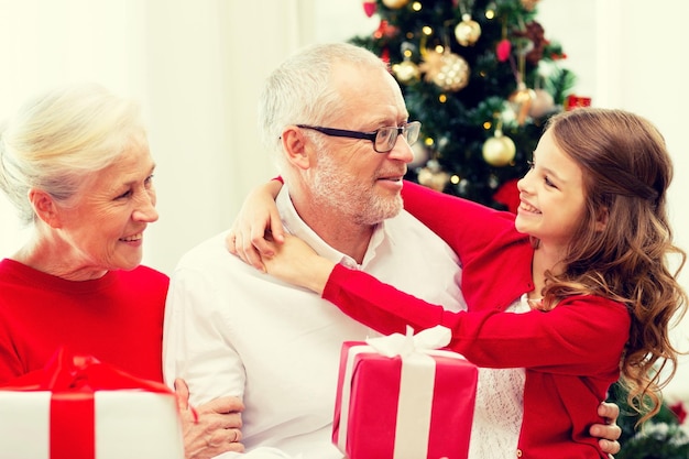 family, holidays, generation, christmas and people concept - smiling grandparents and granddaughter with gift boxes sitting on couch at home