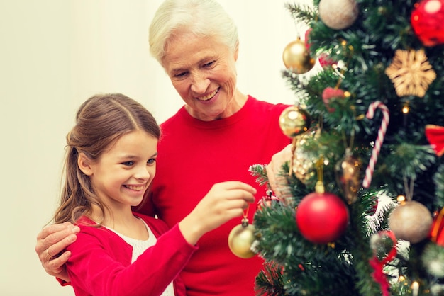 family, holidays, generation and people concept - smiling girl with grandmother decorating christmas tree at home