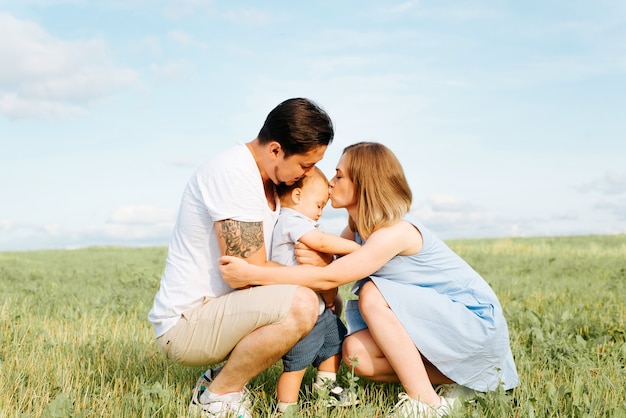 Family hugs and caresses, dad and mom kiss their little child, spending time together, walking, leisure in green field outside. Family portrait outdoors