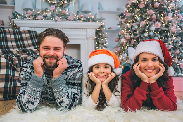 Family look. Young man, woman and daughter lying on floor and look. They pose. people hold hands under chin. They smile.