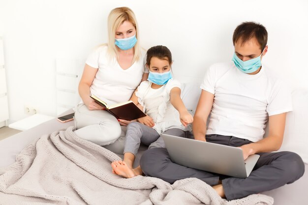 Family in medical face masks lying on a bed with a laptop. Concept of watching video, working together and quarantine during a COVID-19 coronavirus epidemic