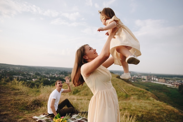 Family, mom, dad, daughter on picnic on a hill in the field. Mom and daughter in yellow identical dresses. Sunflowers.