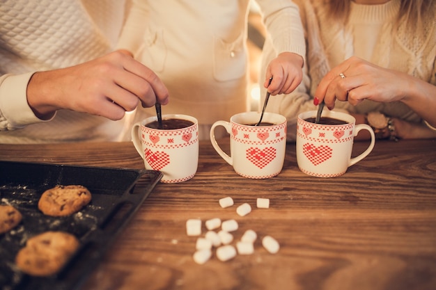 Family mom, dad and daughter in white sweaters cook and drink cocoa with marshmallows