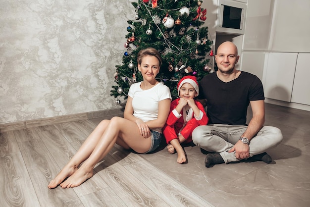 Family mom dad and son in Santa costume sitting under the Christmas tree on the floor in their apartment looking into the camera