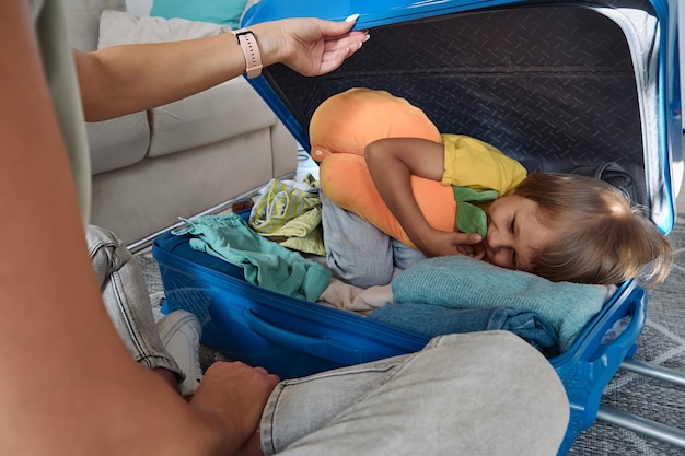 Family mother and child are having fun the child is hiding in a suitcase packing things and travel pillow for a trip