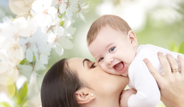 family, motherhood, children, parenthood and people concept - happy mother kissing her baby over cherry blossom background