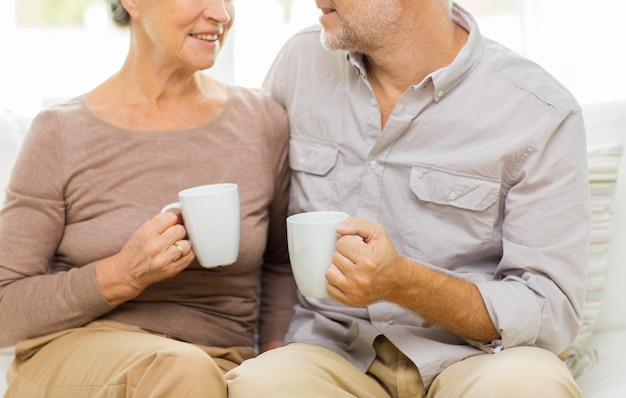 family, relations, age, drinks and people concept - close up of happy senior couple with cups hugging on sofa at home