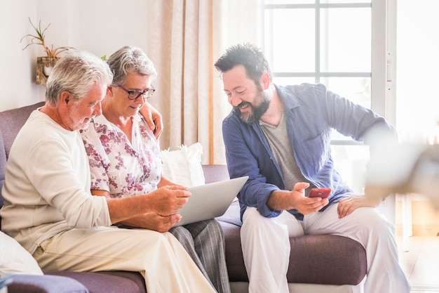 Family sitting together on the sofa using modern device laptop and phone and enjoying connection to web internet Father mother and son smile and enjoy Adult and senior people at home