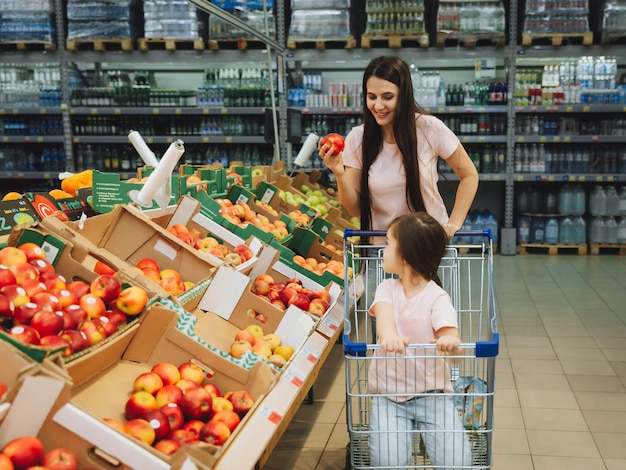 Family in the supermarket young mother and her little daughter are smiling and buying food Healthy food concept mom and daughter buy vegetables and fruits in the supermarket