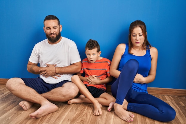 Family of three sitting on the floor at home with hand on stomach because indigestion, painful illness feeling unwell. ache concept.