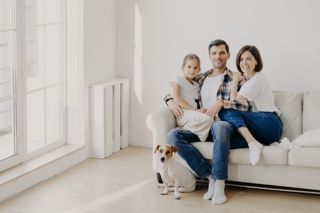 Family, togetherness and relationship concept. Happy man embraces daughter and wife, sit on comfortable white sofa in empty room, their pet sits on floor, make family portrait for long memory