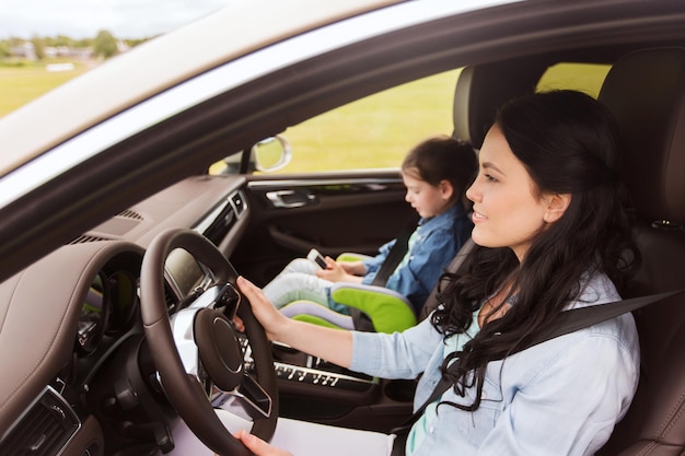 family, transport, road trip, travel and people concept - happy woman and little daughter with smartphone driving in car