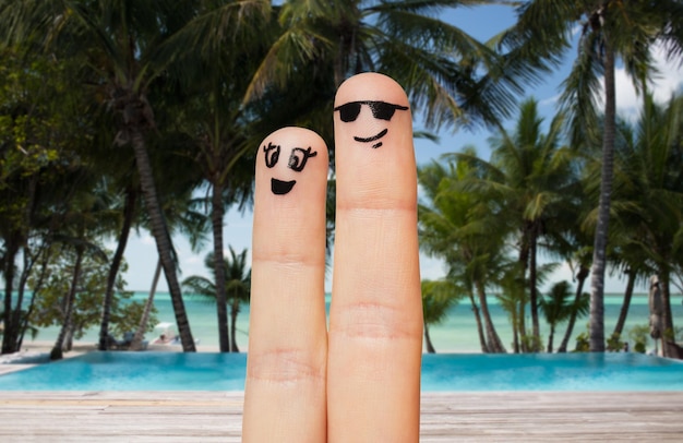 family, travel, summer holidays, tourism and body parts concept - close up of two fingers with smiley faces over exotic tropical beach with palm trees and pool background