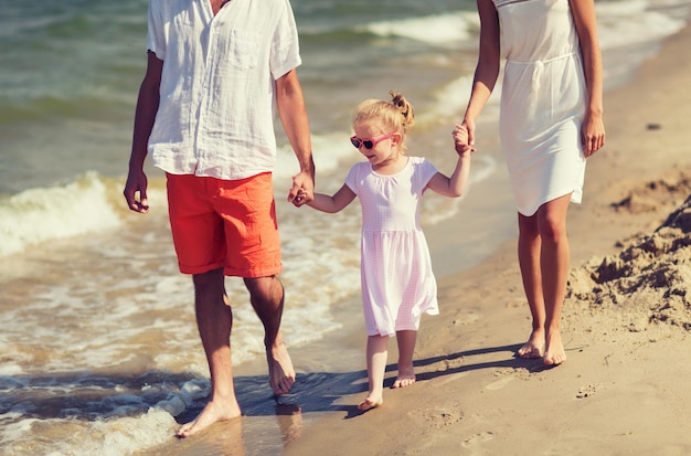 family, vacation, adoption and people concept - happy man, woman and little girl in sunglasses walking on summer beach