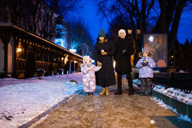 Family walk through the night city in the winter A woman and three children in evening drink hot drink in cups