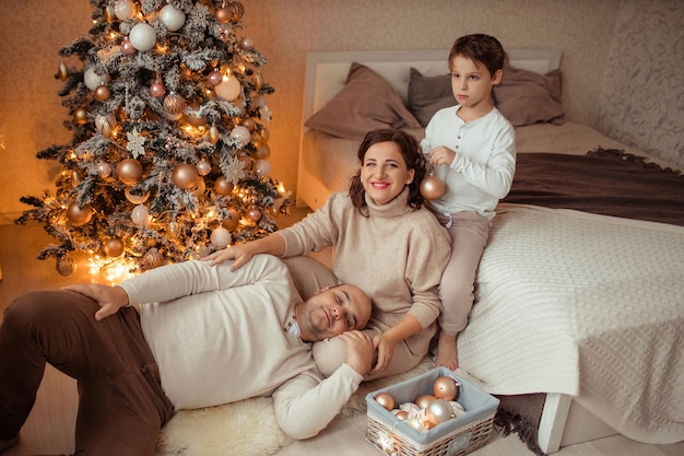 A family with a child at home in the bedroom by the bed near the Christmas tree.