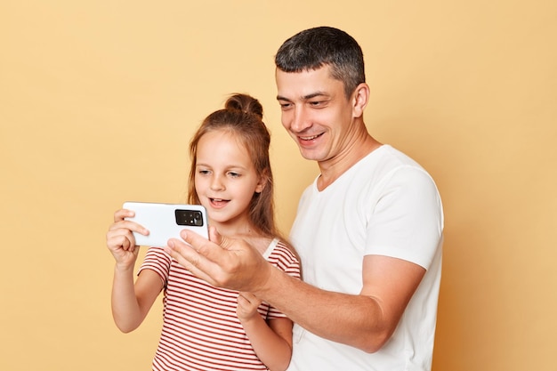 Family with happy facial expression father and daughter wearing casual tshirts standing isolated over beige background using smartphone watching cartoons on phone