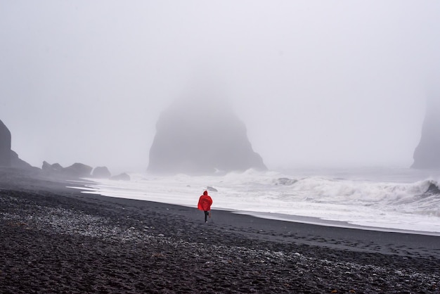 Famous black sand beach Vik in Iceland Person in red raincoat running by the sea shore in the fog