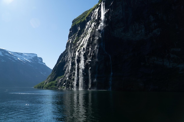 Famous seven sisters waterfall sliding down the cliff into Geirangerfjord on a sunny day with snowcapped mountains in the background