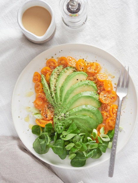 Fanshaped avocado served with yellow cherry tomatoes and green leaves Top view of plate with fork on table next to pepper grinder and dressing in jar