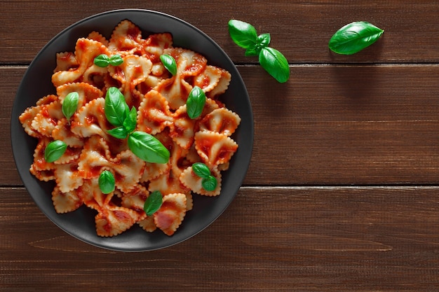 Farfalle cooked pasta with tomato sauce and basil green leaves in gray plate on wooden brown plank tabletop background top view space to copy textxA