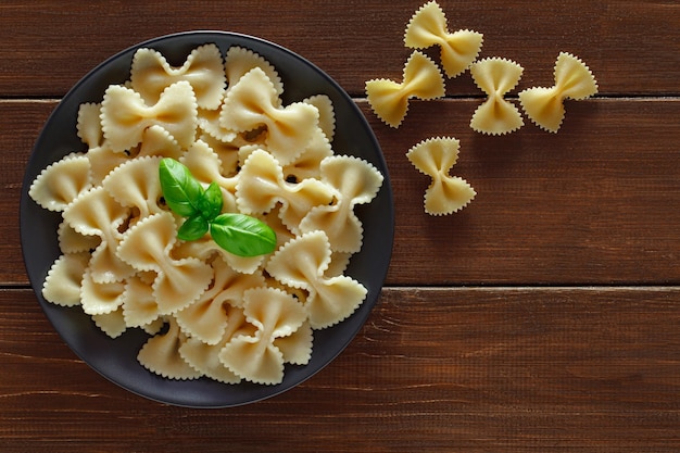 Farfalle cooked and raw pasta with basil green leaves on wooden brown plank tabletop background top view space to copy textxA