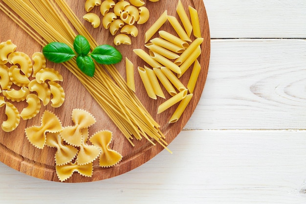 Farfalle spaghetti pipe rigatoni maccheroni penne rigate cresta di gallo raw basil on round wooden plank on white wooden board background top view space to copy textxA