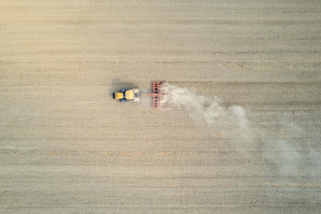 Farmer driving modern yellow crawler tractor harrowing and leveling dry soil for next seeding in rural plain fields aerial drone shot