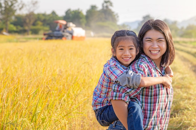 Farmer mother giving daughter piggyback ride at golden-yellow rice field with combine tractor