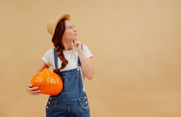 Farmer woman in overalls holds a harvest large pumpkin looking thoughtfully to the right side