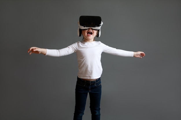 Fascinated female child in white long sleeve shirt wearing virtual reality glasses over grey surface.