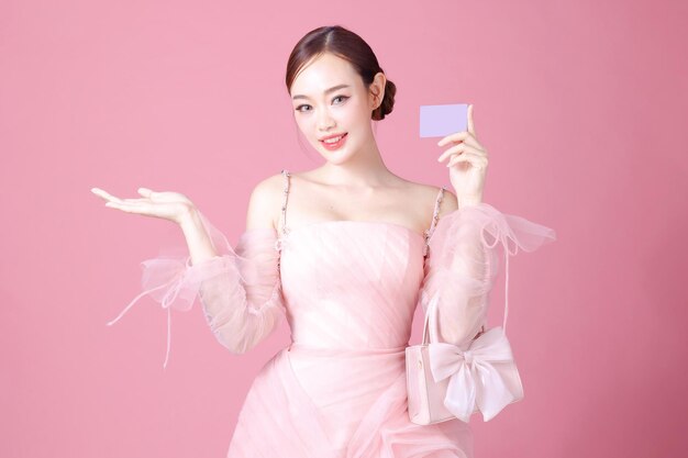 Fascinating fun joyful young woman of Asian wears sweet pink flower dress hold in hand credit bank card cover isolated on plain pastel light pink background Beautiful lady portrait in studio