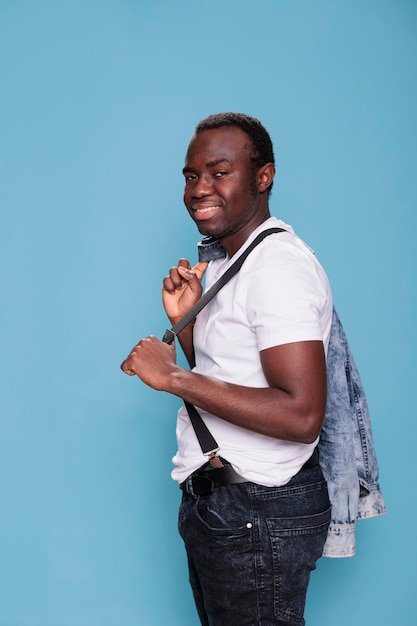 Fashion african ethnicity man holding denim jacket on back while smiling at camera and standing on blue background. Cheerful and handsome young person wearing pulling suspenders. Studio shot