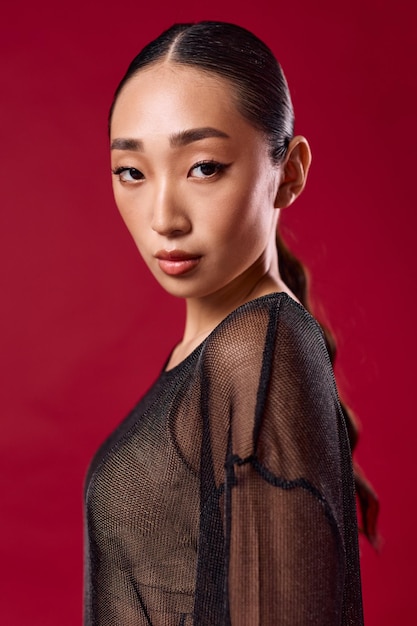 Photo fashion asian woman in sheer black top poses against red background with hair in ponytail and eyes