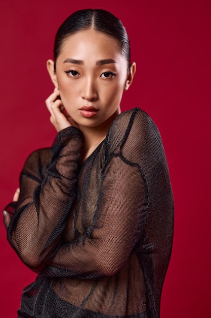 Photo fashion asian woman in sheer top and black skirt posing against red background with hands on hips