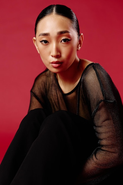 Fashion asian woman sitting in sheer top and black pants on red background with eyes closed