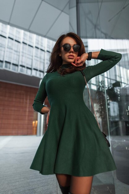 Photo fashion beautiful chic lady with stylish sunglasses in a fashionable green dress with handbag walks in the city near the modern business glass office building