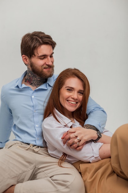 Fashion beautiful happy couple sitting in the studio Fashionable happy redhead model woman with cute smile and handsome brutal hipster man with tattoo and beard