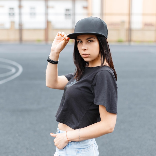 Photo fashion beautiful young hipster girl in a black tshirt puts on a black baseball cap at the stadium