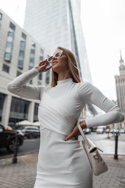 Fashion beautiful young woman in a white fashion dress with a handbag puts on white stylish sunglasses and walks in the city Urban fashion vogue and beauty