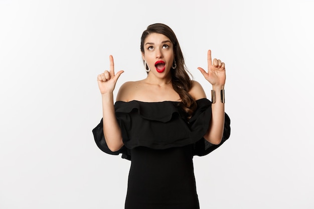 Fashion and beauty. Amused woman in black dress looking and pointing fingers up at promo offer, standing over white background