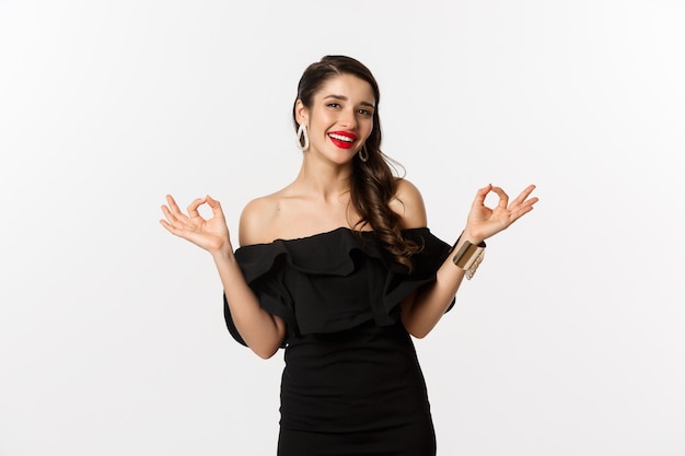 Fashion and beauty. Attractive brunette woman in black dress, showing okay signs and smiling satisfied, approve and recommend, standing over white background.