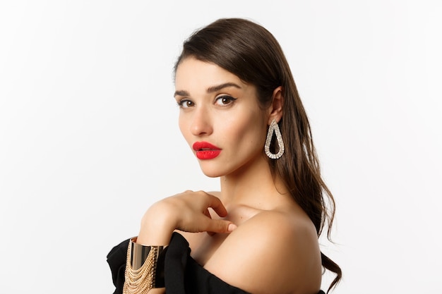 Fashion and beauty concept. Close-up of elegant woman with red lips, makeup and earrings, looking at camera self-assured, standing over white background