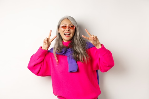 Fashion and beauty concept. Image of stylish asian senior woman in sunglasses smiling, showing peace signs and looking happy, standing over white background